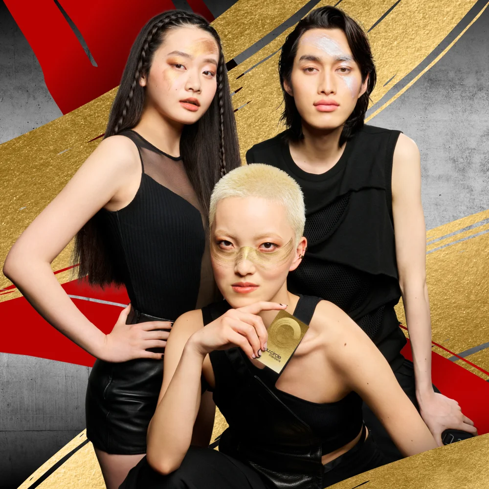alt="Three models in black outfits with bold editorial gold makeup posing against a dramatic gold, red and grey brushstroke background, the centre model holding a Shu Uemura Circle rewards card."