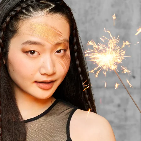 alt="Model with braided hair and bold gold editorial eye makeup holding a sparkling firework against a grey background.alt="Model with braided hair and bold gold editorial eye makeup holding a sparkling firework against a grey background."