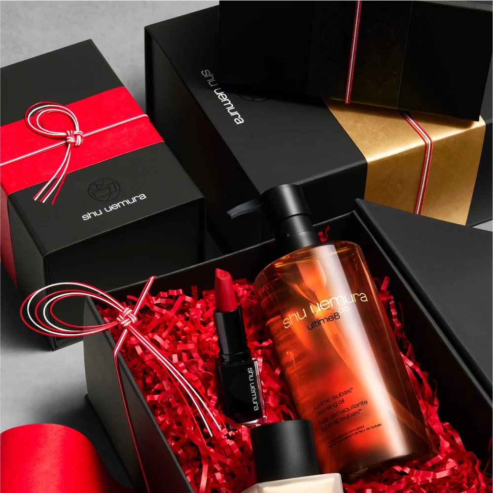 alt="Shu Uemura gift set in an open black box with red paper fill, containing the Ultime8∞ cleansing oil bottle and a red lipstick, surrounded by additional black and gold gift boxes tied with red and white Japanese mizuhiki cord."