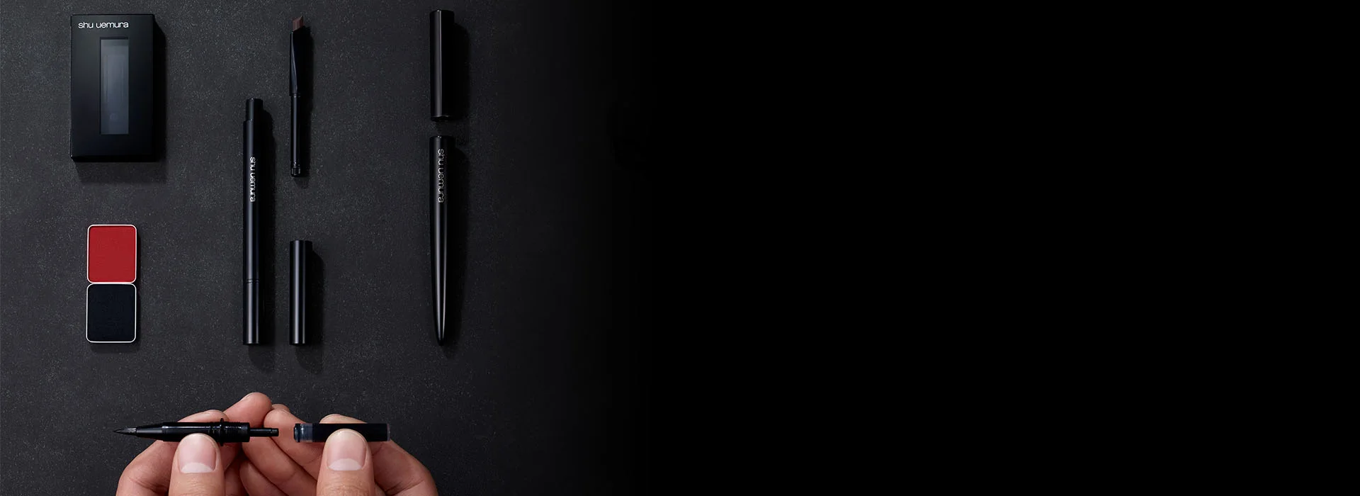 alt="A collection of Shu Uemura makeup, including an eyeshadow duo, eyebrow pencils, and brushes, laid out on a dark surface while a hand assembles a precision tool."