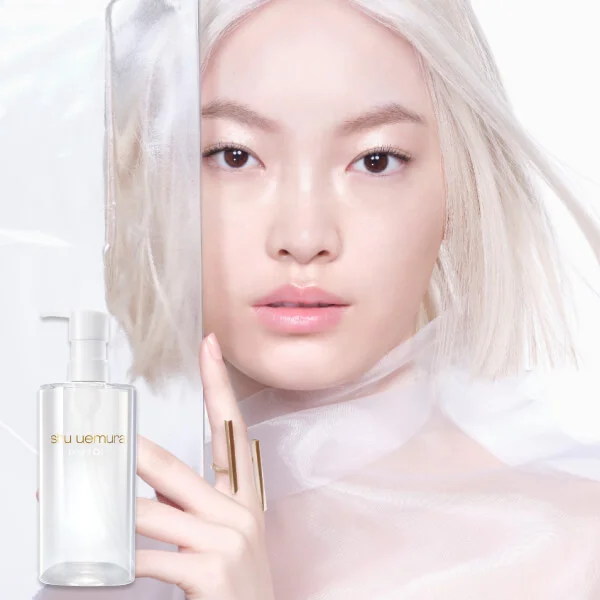 alt="Model with platinum blonde hair and luminous bare skin holding a Shu Uemura clear cleansing oil pump bottle against an all-white background."